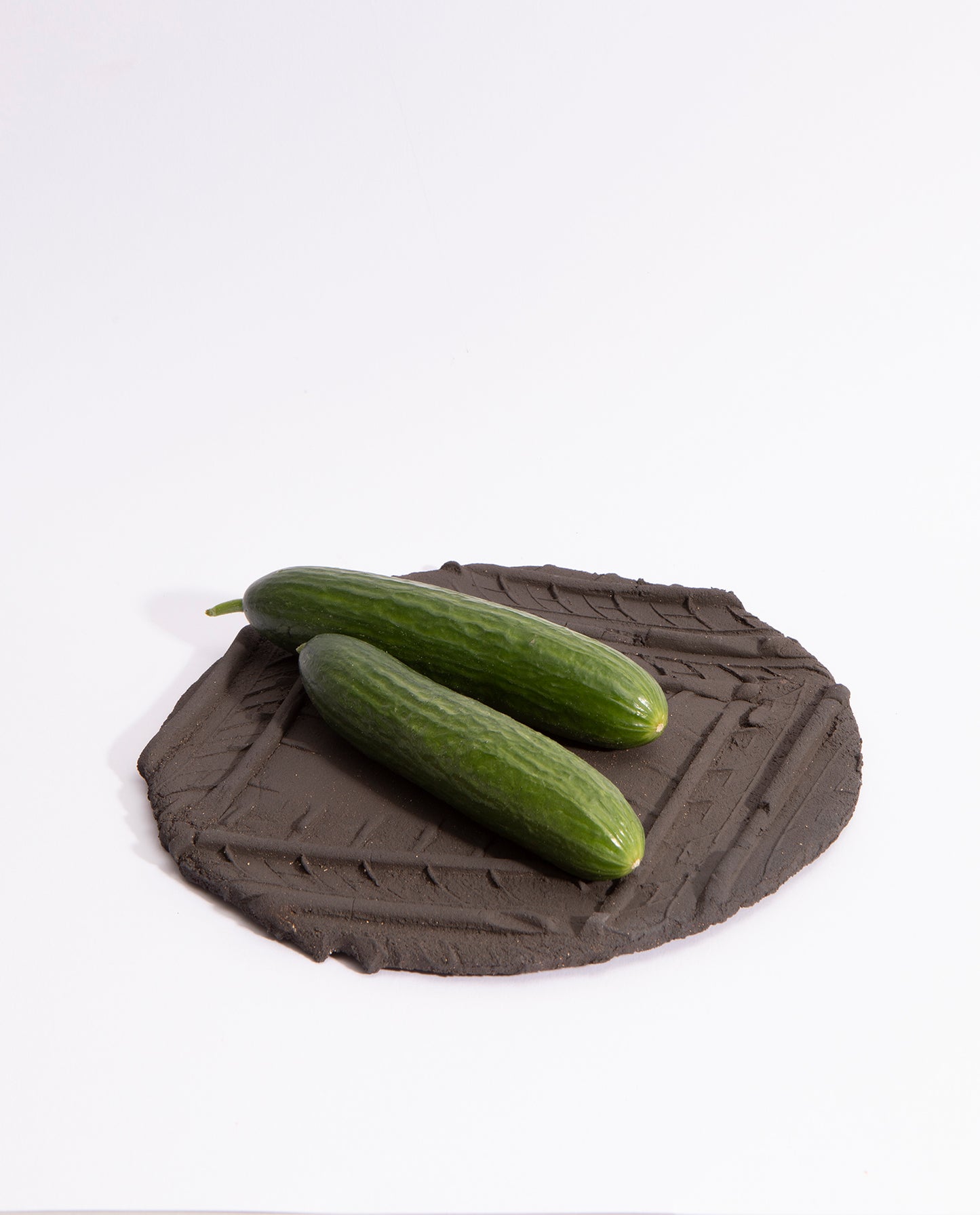 Assiette en grès noir brut, avec quatre empreintes de pneu qui forme un carré sur l'assiette. Deux concombres sont dessus.