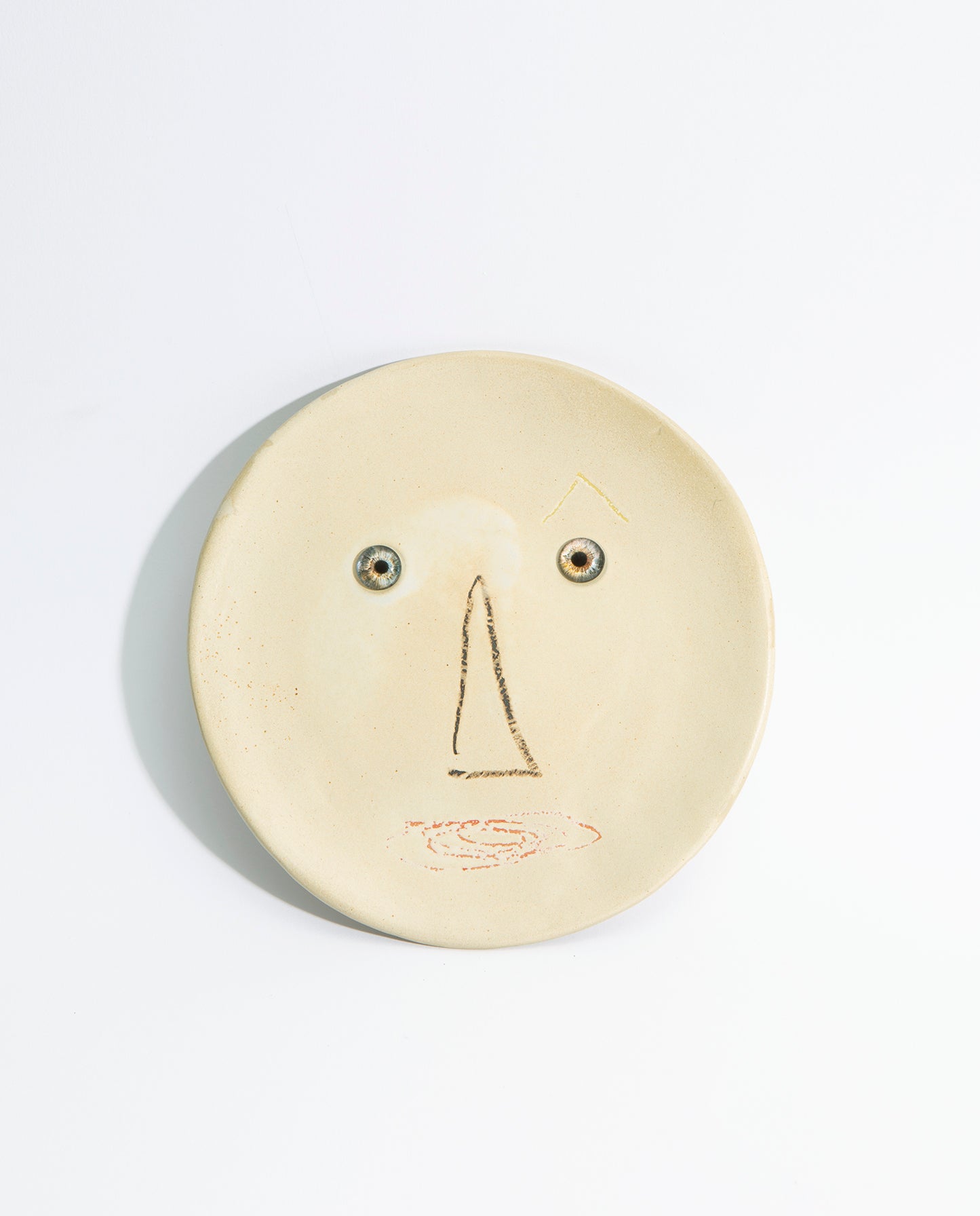 Assiette en grès beige, représentant un visage en dessin avec deux yeux en verre.